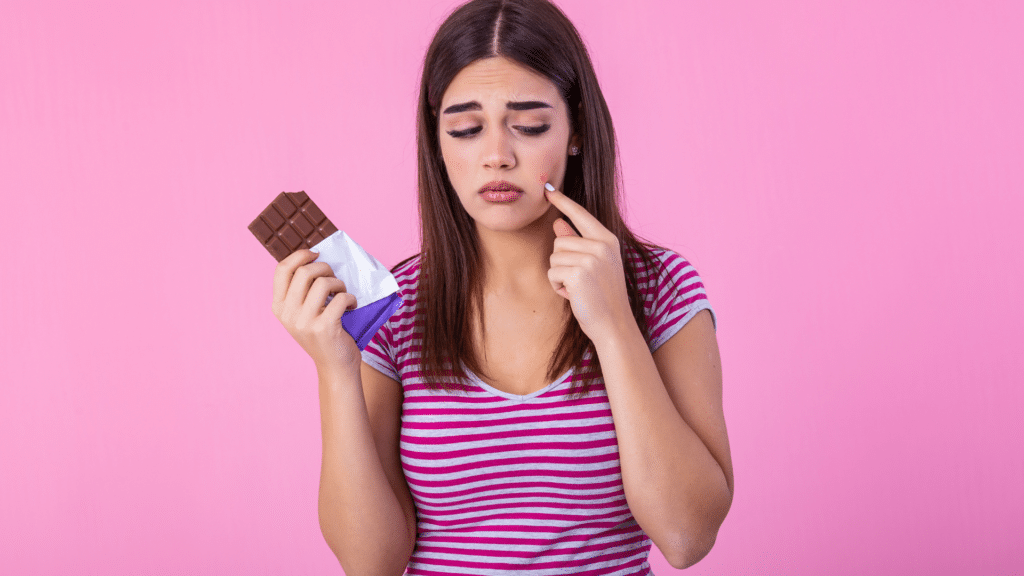 girl holding a chocolate bar pointing at an acne blemish on her face
